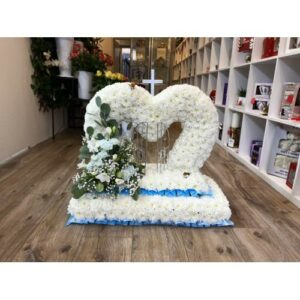 Heart-shaped floral arrangement with a combination of white and yellow flowers on a blue base, displayed indoors, symbolizing the Heart Gates of Heaven.