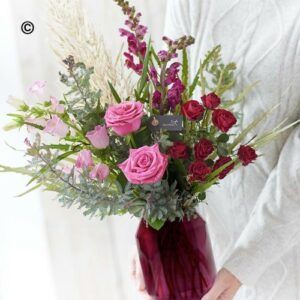 A person holds a bouquet of red and pink roses with mixed foliage in a Valentines Vase.