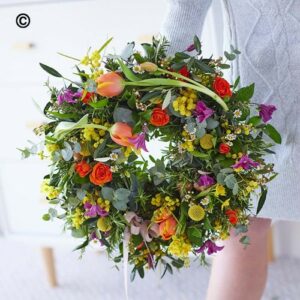 A person holding a colorful Spring Wreath featuring a mix of flowers and greenery.