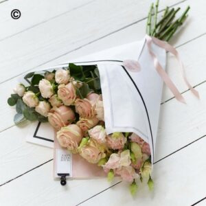 A fresh bouquet of pink roses wrapped in paper with a pink ribbon next to a white tube on a wooden surface is Simply Beautiful Deluxe.