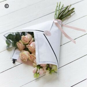 A Simply Beautiful bouquet of pink roses wrapped in paper with a pink ribbon on a white wooden surface.