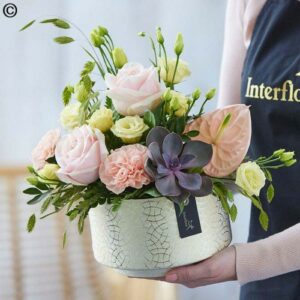 A person holding a Seasonal Flowers arrangement featuring pink roses, greenery, and a purple succulent in a cracked-patterned ceramic pot.