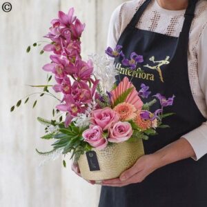 A person holding a vibrant Magnificent Orchid arrangement, including pink roses and purple orchids, arranged in a textured gold vase.