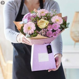 Person presenting a Sunflower Surprise Handtied Bouquet of flowers wrapped with brown paper in a purple box.