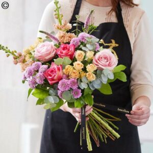 A person wearing an apron is holding a Handtied Bouquet Countryside of mixed flowers, which includes pink roses and purple accents.