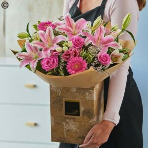 Person holding a bouquet of pink flowers, including Hand Tie Roses and Lilys, wrapped in decorative paper.