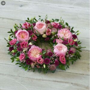 A deluxe wreath of All Pink Deluxe Wreath roses and assorted purple flowers on a wooden surface.