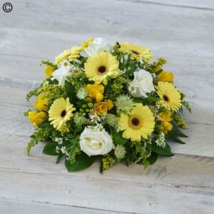 A bright Yellow Posy and white floral arrangement with gerberas, roses, and mixed greenery on a wooden surface.