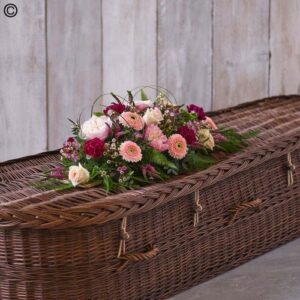 Petite wicker casket adorned with a Petite Casket Spray against a wooden backdrop.