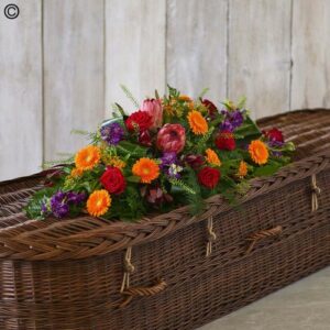 A wicker casket adorned with a Tropical Casket Spray against a wooden background.