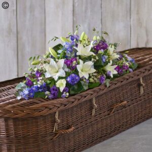A wicker casket adorned with a White and Purple Casket Spray of white lilies, purple flowers, and green foliage.