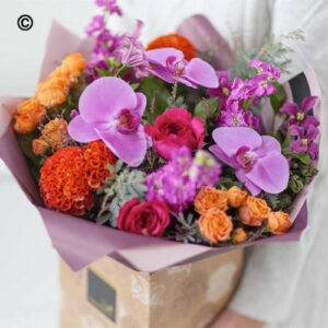 A Orchid Valentines Handtie bouquet of vibrant purple orchids, orange roses, and assorted flowers wrapped in purple paper.
