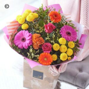 A colorful bouquet of Vivid Handtie including yellow, pink, and orange blooms, handtied by a florist, held by a person standing behind it.