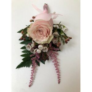 A single pink rose surrounded by small white flowers and green leaves, arranged as a Wedding boutonniere or corsage with pink ribbons against a white background.